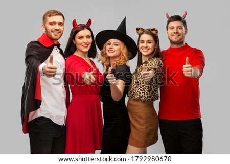 friendship, holiday and people concept - group of happy smiling friends in halloween costumes of vampire, witch, devil and cheetah showing thumbs up over grey background