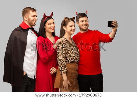friendship, holiday and people concept - group of happy smiling friends in halloween costumes of vampire, devil and cheetah taking selfie by smartphone over grey background