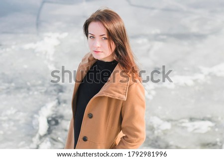 Portrait of a beautiful woman on the waterfront. Thoughtful young woman in brown coat on the river bank, photo taken in winter in January