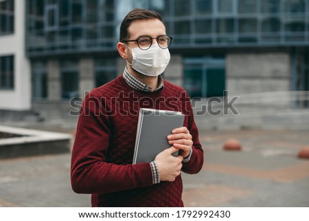 Portrait of a man in glasses, in a medical mask, with books, notebook in his hands, on the background of a modern building. Education at the university during quarantine. Coronavirus, protection. Royalty-Free Stock Photo #1792992430