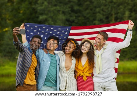 Waist up portrait of multi-ethnic group of friends holding American flag and taking selfie outdoors while enjoying party in Summer, copy space