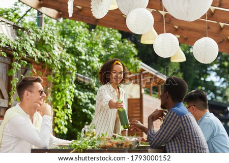 Multi-ethnic group of people enjoying dinner at outdoor terrace in Summer, focus on smiling African-American woman handing bottle of wine across table, copy space