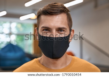 Back to office. Close up portrait of young man, male office worker wearing black protective mask and looking at camera, standing in the modern office