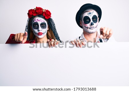 Couple wearing day of the dead costume holding blank empty banner pointing with finger to the camera and to you, confident gesture looking serious 