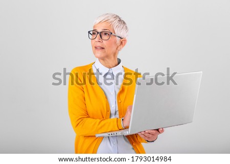 Photo of a thinking mature business woman isolated over grey background holding laptop computer. Image of confused senior woman using laptop computer. Looking at laptop while holding face with hands.