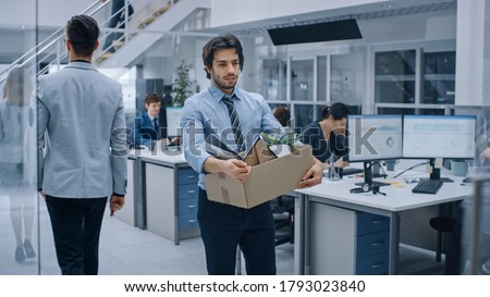 Sad Fired / Let Go Office Worker Packs His Belongings into Cardboard Box and Leaves Office. Workforce Reduction, Downsizing, Reorganization, Restructuring, Outsourcing. Shot with Dark Ambient Royalty-Free Stock Photo #1793023840