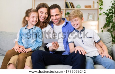 Family Of Four Using Mobile Phone Watching Cartoons Together Sitting On Sofa At Home. Weekend Leisure