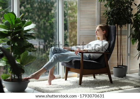 Young woman at home sitting on modern chair near window relaxing in living room Royalty-Free Stock Photo #1793070631
