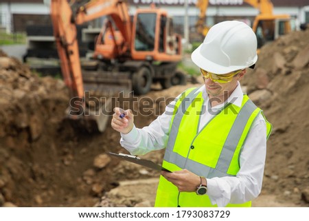 A man in construction clothes with a white helmet in a white shirt on the background of an excavator. Engineering concept on the construction site next to the excavation equipment