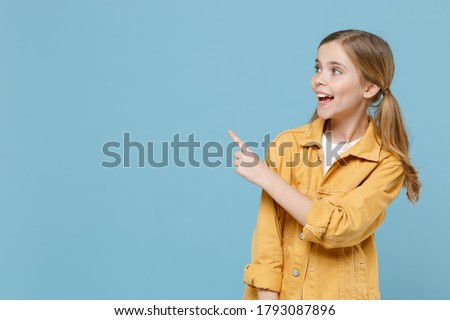 Funny little blonde kid girl 12-13 years old in yellow jacket isolated on blue background children studio portrait. Childhood lifestyle concept. Mock up copy space. Pointing index finger aside up