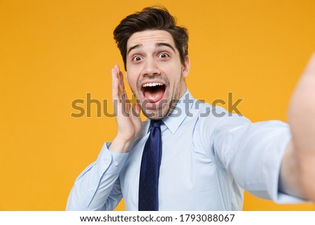 Close up of shocked young business man in classic blue shirt tie isolated on yellow background studio. Achievement career wealth business concept. Doing selfie shot on mobile phone put hand on cheek