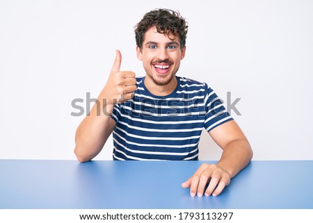 Young caucasian man with curly hair wearing casual clothes sitting on the table smiling happy and positive, thumb up doing excellent and approval sign 