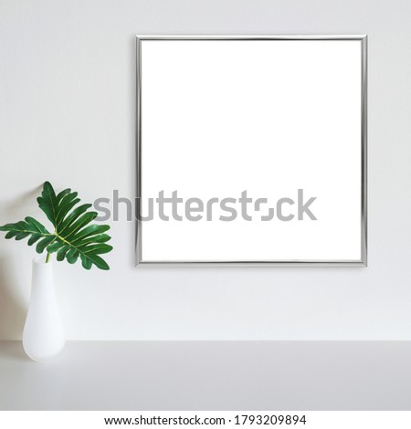 Silver frame mockup on white wall with philodendron leaf in vase on white surface. Copy space.