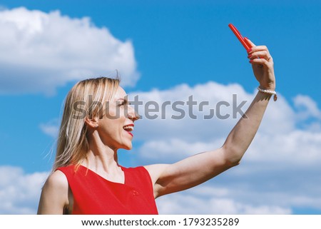 Blonde woman with red dress taking selfie with cell phone in blue with white clouds sky background with copy space during sunny summer day