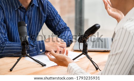 Asian woman radio hosts gesturing to microphone while interviewing a man guest in a studio while recording podcast for online show in studio together.