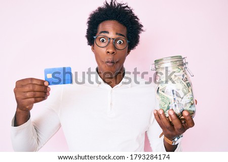 Handsome african american man with afro hair holding credit car and jar with dollars making fish face with mouth and squinting eyes, crazy and comical. 
