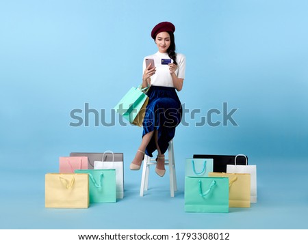 Beautiful Asian woman shopper sitting and carrying shopping bags with using credit card and mobile phone in hands on blue background. Royalty-Free Stock Photo #1793308012