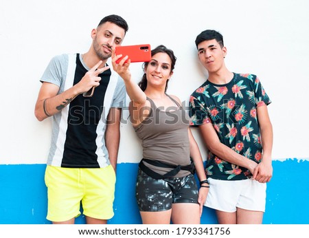 three young people, in summer clothes, on a white and blue wall, using their smartphones