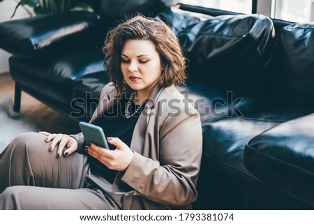 Caucasian brunette woman sitting near sofa in living room and using phone for freelance job.
