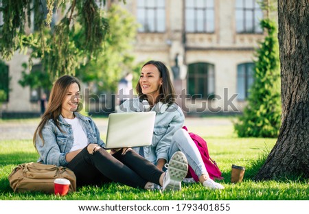 Two happy beautiful young student girl friends in casual denim clothes are relaxing in college park with laptop and smartphone on university background and drinking coffee.