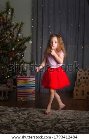 Little girl in a red skirt is dancing for the New Year. The girl at the Christmas tree is having fun at home.