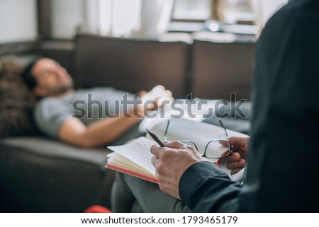 Psychotherapist and patient at an appointment in the office. Doctor's hands with notepad close-up. The doctor records the client's history. A male patient lies on a sofa in a large, bright office Royalty-Free Stock Photo #1793465179