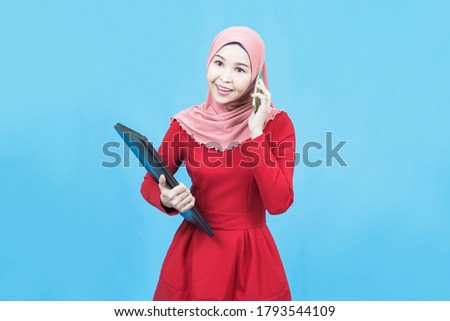Portrait of young beautiful asian business muslim woman in red traditional dress holding laptop and talking on the phone isolated on blue background