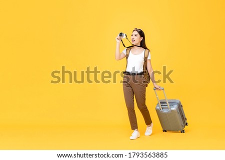 Full length travel portrait of excited young tourist Asian woman walking while taking photo with camera in isolated studio yellow background