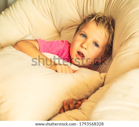 Blond surprised boy with open mouth in pink t-shirt sit with pillow in round fashionable chair in living room in a house indoor. Family relationship, childhood problems behaviour, education psychology