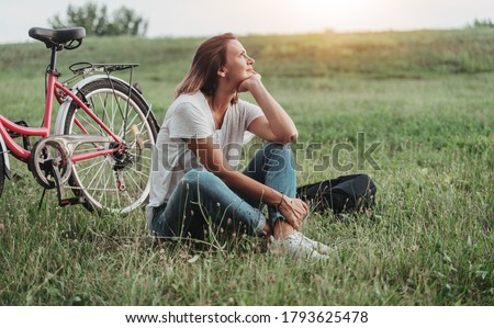 Beautiful smiling mature woman sitting next to bike on the grass on a green field. Summer Country Vacation and Adventure Concept Royalty-Free Stock Photo #1793625478