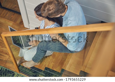 Back view portrait of cheerful female and handsome man wearing casual clothing sitting on the wooden stairs while looking at the screen of notebook