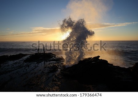 Water is shot up and out of a blowhole in the rocks in front of the early morning sunrise Royalty-Free Stock Photo #179366474
