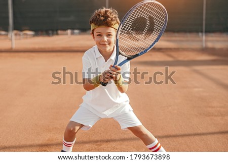 Positive active school aged boy with racket looking at camera while playing tennis on court Royalty-Free Stock Photo #1793673898