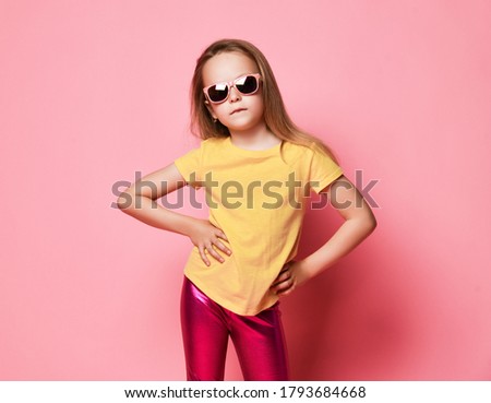 Cool stylish kid girl in modern sunglasses, yellow t-shirt and pink glossy leggings stands posing as a model with her hands on her waist over pastel pink background