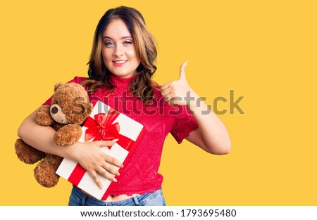 Young beautiful caucasian woman holding gift and teddy bear smiling happy and positive, thumb up doing excellent and approval sign 
