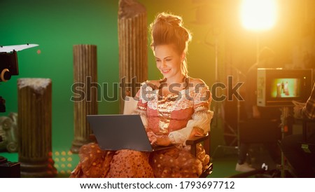 On Period Costume Drama Film Set: Beautiful Smiling Actress Wearing Renaissance Dress, Sitting on Chair Using Laptop Computer with Green Screen in Background. Studio Shooting Historical Blockbuster