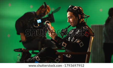Handsome Smiling Actor Wearing Motion Capture Suit and Head Rig having Lunch Break, Sitting on Chair, Uses Smartphone. Studio High Budget Movie. On Film Studio Period Costume Drama Film Set