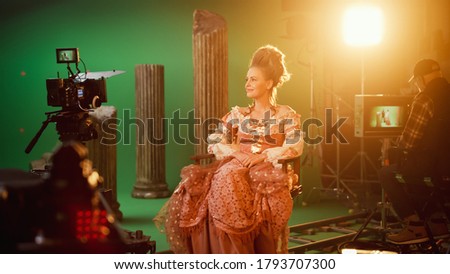 On Period Costume Drama Film Set: Beautiful Smiling Actress Wearing Renaissance Dress, Sitting on a Chair Happily with Green Screen in the Background. Studio Shooting History Blockbuster