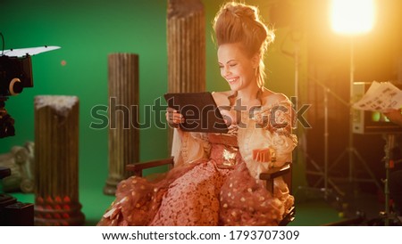 On Period Costume Drama Film Set: Beautiful Smiling Actress Wearing Renaissance Dress, Sitting on Chair Using Digital Tablet Computer with Green Screen in Background. High Budget Period Drama.