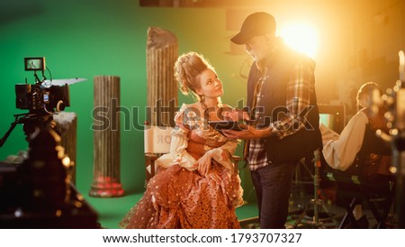 On Period Costume Drama Film Set: Beautiful Smiling Actress Wearing Renaissance Dress Sitting on a Chair Listens to Movie Director Explaining to Her Scene Context. High Budget Period Drama.