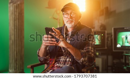 Prominent Successful Senior Director Sitting in a Chair on a Break Using Smartphone and Looking at Camera. On the Studio Film Set with High-End Equipment Professional Crew Shooting High Budget Movie