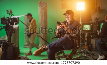 Prominent Successful Director Sitting in a Chair on a Break Using Smartphone. On the Studio Film Set with High-End Equipment Professional Crew Shooting High Budget Movie.