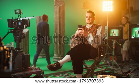 On Period Drama Film Set: Handsome Actor Wearing Historical Costume Sitting in a Chair on a Break Using Smartphone. In the Studio Professional Crew Shooting High Budget Movie