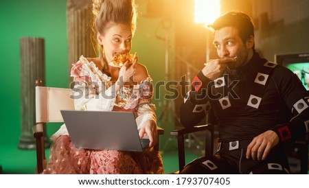 Beautiful Smiling Actress Wearing Renaissance Dress and Actor Wearing Motion Capture Suit having Lunch Break, Sitting on Chairs, Use Laptop and Talk. Studio High Budget Movie. Costume Drama Film Set