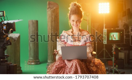 Beautiful Smiling Actress Wearing Renaissance Dress, Sitting on a Chair Using Laptop Computer with Green Screen in the Background. On Film Studio Period Costume Drama Film Set