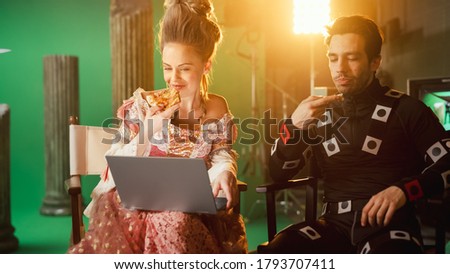 Beautiful Smiling Actress Wearing Renaissance Dress and Actor Wearing Motion Capture Suit having Lunch Break, Sitting on Chairs, Use Laptop and Talk. Studio High Budget Movie. Costume Drama Film Set