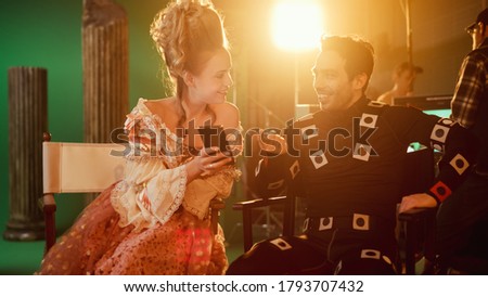 Beautiful Smiling Actress Wearing Renaissance Dress and Actor Wearing Motion Capture Suit Sitting on Chairs Share Social Media Posts via Smartphone. On Film Studio Period Costume Drama Film Set