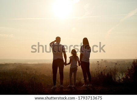 Silhouette of a family - father, mother and daughter standing on the hill with their backs to the camera and looking to the horizon on the sunset