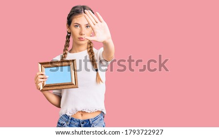 Beautiful caucasian woman with blonde hair holding empty frame with open hand doing stop sign with serious and confident expression, defense gesture 