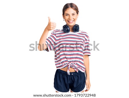 Beautiful caucasian woman with blonde hair wearing gym clothes and using headphones smiling happy and positive, thumb up doing excellent and approval sign 